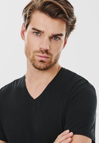 Czarny t-shirt z dekoltem w serek, wykonany z gładkiego materiału, z krótkim rękawem i luźnym krojem. Model pozuje ze skrzyżowanymi ramionami na jednolitym tle.