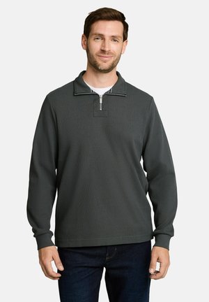 Homme portant un pull-over gris foncé à manches longues avec une fermeture éclair quart de zip et une texture côtelée, ainsi qu’un jean bleu foncé, debout sur un fond uni.