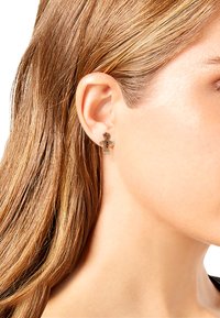 Pendientes en forma de hombre de jengibre en tono dorado con acentos rojos, que presentan un acabado pulido y un detalle intrincado, llevados en la oreja de un modelo.