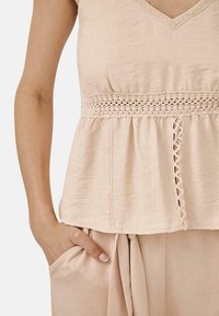 Blusa de color durazno con escote en V, tela texturada, detalles de encaje en la cintura y corte holgado. Incluye bolsillos laterales.