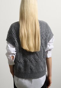 Persoon met lang blond haar, gekleed in een grijze gebreide mouwloze trui met textuur over een witte lange mouw shirt, staande tegen een plain muur.