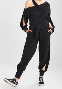 Svart kort sweatshirt med asymmetrisk halsringning och strukturerade ärmar, tillsammans med svarta joggers med bälte i midjan och sidoslitsar.