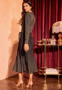 Metallisch goldenes, plissiertes Kleid mit einem Bindegürtel, langen Ärmeln und midi-langer Länge, kombiniert mit goldenen Riemchenheels vor einem burgunderfarbenen Hintergrund.