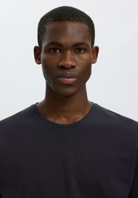 Joven hombre negro con cabello corto, vistiendo una camiseta oscura lisa con cuello redondo, mirando al frente sobre un fondo gris claro.