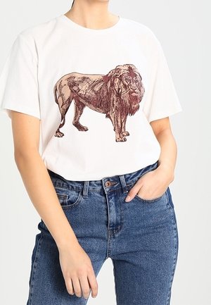 Persoon die een wit T-shirt draagt met een gedetailleerde bruine leeuwgrafiek, gecombineerd met hoog getailleerde blauwe spijkerbroek, één hand in de zak.