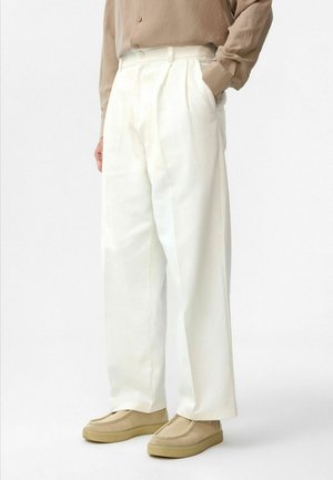 Witte, wijde broek met plooien, gemaakt van een lichte stof. Gecombineerd met beige instappers en een taupe blouse met knopen.