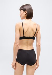 Γυναίκα εμφανίζεται από πίσω φοράει μαύρο σουτιέν με ρυθμιζόμενες τιράντες και μαύρο σλιπ με ψηλή μέση, σε απλό φωτεινό φόντο.