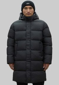 Abrigo de plumas largo y negro con capucha, diseño acolchado rectangular, cierre delantero con cremallera y puños elásticos. Llevado con un gorro de punto negro.