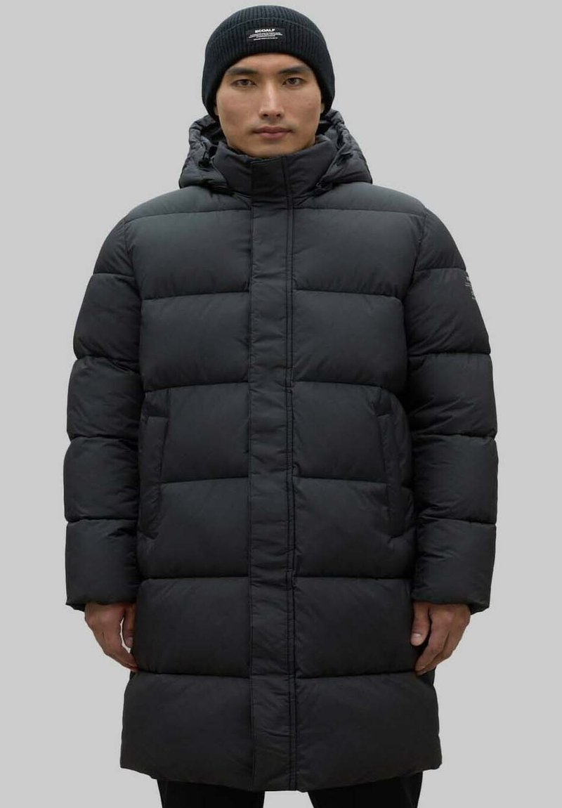 Abrigo de plumas largo y negro con capucha, diseño acolchado rectangular, cierre delantero con cremallera y puños elásticos. Llevado con un gorro de punto negro.