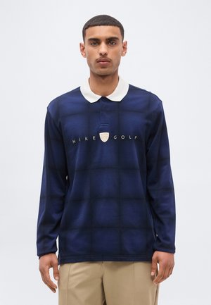 Muž v tmavomodrom dlhom rukáve Nike Golf polo s bielym golierom a béžovými nohavicami, stojaci pred jednoduchým svetlým pozadím.