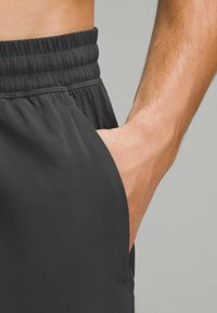 Mörka träningsshorts med elastisk midja och sidofickor. Slät textur med en solid svart färg. Närbild av en hand som når in i fickan.