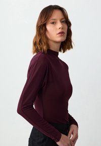 Burgunderfarbene Langarm-Bluse mit hohem Kragen, glatter Textur, figurbetontem Design und dezentem Glanz, kombiniert mit dunklen Hosen.
