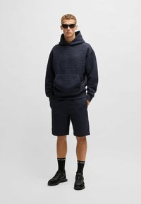 Navy hoodie with hexagonal patterns and kangaroo pocket, paired with matching shorts. Worn with black sunglasses, socks, and sneakers.