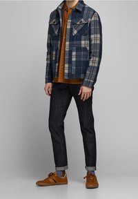 Kostkovaná bunda v modro-bežové vlně s knoflíkovou přední částí, kombinovaná s hnědou košili a tmavými džíny. Hnědé boty outfit dokonal.