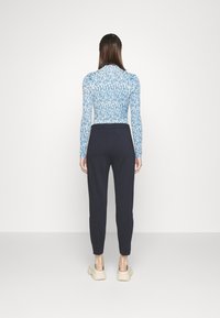 Kvinna som bär en långärmad topp i blått och vitt mönster, svarta åtsittande byxor och beiga slip-on skor, stående mot en enkel vit bakgrund.