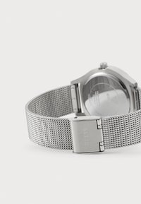 Zegarek na srebrnej siateczkowej bransolecie z okrągłą kopertą. Posiada zapięcie z grawerunkiem "GUESS". Minimalistyczny design z gładkim metalicznym wykończeniem.
