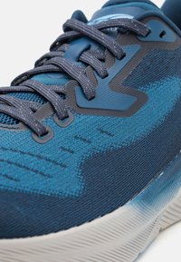 Close-up of blue athletic shoe with textured mesh fabric, dark blue laces, and white cushioned sole.