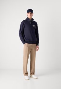 Hanorac bleumarin cu fermoar și logo, purtat cu pantaloni bej. Tenisi albi completează ținuta. Material fin, croială relaxată.