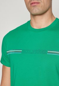 Zielona koszulka bawełniana z okrągłym dekoltem i krótkimi rękawami. Z przodu znajduje się napis "HILFIGER" z kolorowymi paskami akcentującymi powyżej.