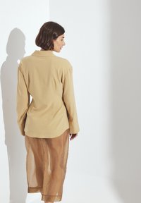 Beige long-sleeve shirt with a collared neckline, paired with sheer brown pants. Smooth fabric with a relaxed fit and rolled cuffs.