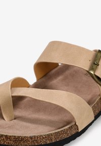 Sandal beige à brides croisées avec une semelle en liège et une surface texturée, dotée d'une accent en boucle métallique et d'une semelle en caoutchouc noir.