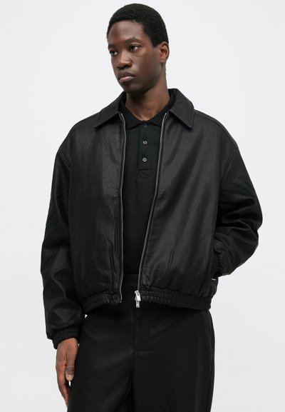 Jonge man in een zwart gestructureerd zip-up jack over een zwart button-down shirt, staand met één hand in zijn zak tegen een eenvoudige achtergrond.