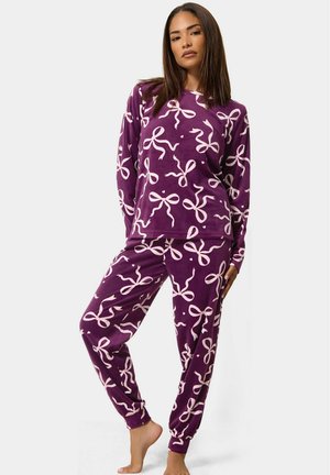 Femme portant un haut à manches longues violet et un pantalon avec un motif de nœuds rose clair, debout pieds nus sur un fond clair uni.