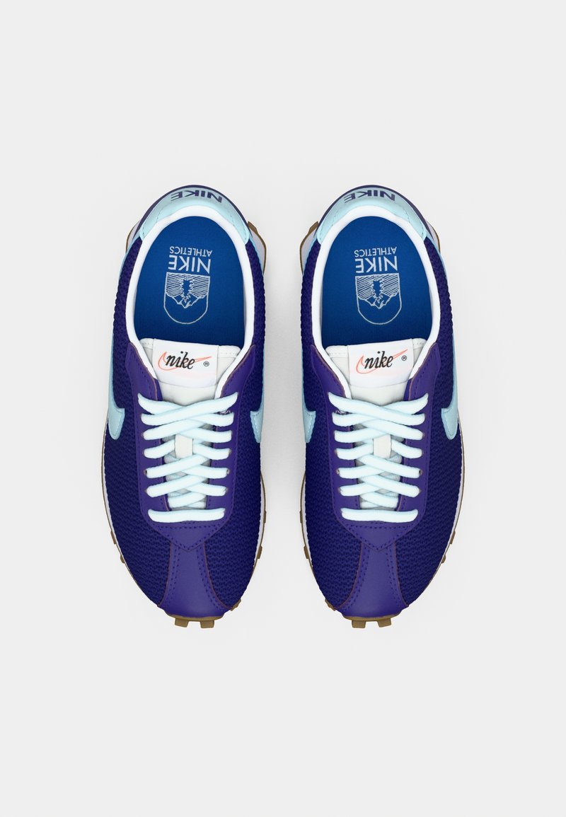 Un par de zapatillas de correr Nike en púrpura y azul claro con cordones blancos y plantillas azules que presentan el logo de Nike Athletics, vistas desde arriba.