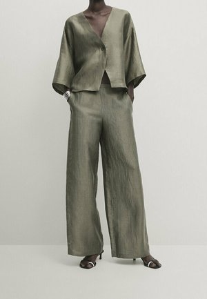 Femme portant un pantalon ample en lin vert olive et un haut portefeuille à manches larges assorti avec des talons ouverts noirs.