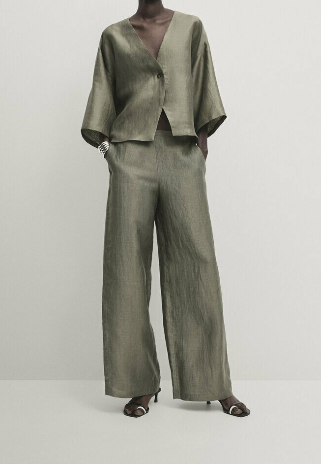 Femme portant un pantalon ample en lin vert olive et un haut portefeuille à manches larges assorti avec des talons ouverts noirs.