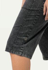 Musta farkkutakki, jossa on haalistunut kuvio, sivusaumat, metallinen nappi ja rento muotoilu, joka päättyy juuri polven ylle.