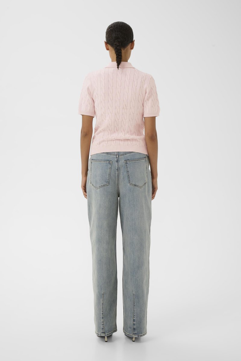 Femme aux cheveux tressés, de dos, portant un pull rose clair à manches courtes et un jean bleu délavé et ample.
