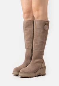 Brown suede knee-high boots with a wedge heel, featuring a side zipper and a round logo accent. Textured surface and contrasting sole.