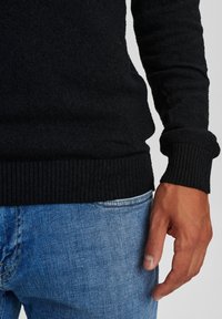 Person som bär en svart stickad tröja och blå jeans, med högerhanden avslappnat vid höfterna mot en ljus bakgrund.