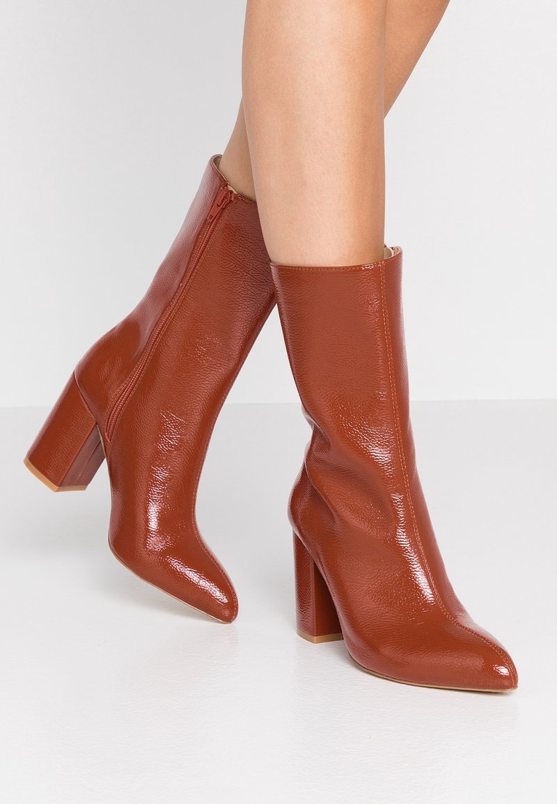 Bottes de cheville marron, brillantes, avec un bout pointu et un talon épais. Texture lisse, tige haute avec un détail de fermeture éclair sur le côté.
