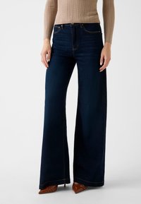 Jean en denim foncé à jambes larges avec une taille haute, des coutures contrastantes et une finition nette à l'ourlet, associé à des chaussures pointues marron.