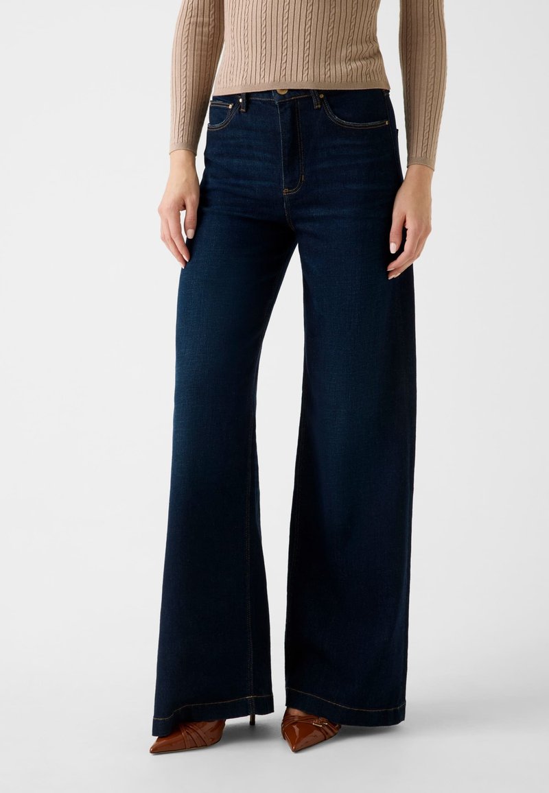 Jean en denim foncé à jambes larges avec une taille haute, des coutures contrastantes et une finition nette à l'ourlet, associé à des chaussures pointues marron.