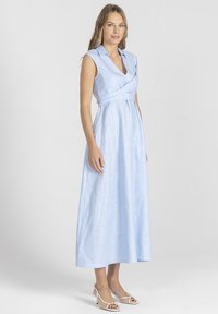 Jasnoniebieska, bezrękawnikowa sukienka z dekoltem w kształcie litery V i zapinaniem w talii, uszyta z gładkiego materiału; zestawiona z beżowymi sandałami na obcasie.