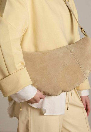 Beige suède schoudertas met een gebogen vorm, voorzien van stiksels en een vergulde hardware band. Zachte textuur, minimalistisch ontwerp.