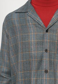 Hallide mustritav hall särk punase kudumisega kõrge kaelusega kampsuni peal, keskendudes kaeluse ja ülemise rinna piirkonnale.