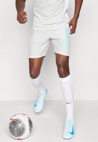 Lys grå treningsshorts med en turkise logo, kombinert med hvite sokker og lys blå fotballsko. En svart-hvit fotball er i nærheten.
