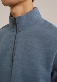 Blå sweatshirt med hög krage och halvdragkedja, gjord av mjukt tyg. Slät textur med ett subtilt melerat mönster.