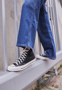 Baskets montantes noires avec semelles en caoutchouc blanches et lacets blancs, portées avec un jean large en denim bleu, posées sur une rambarde gris clair.