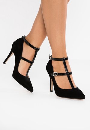 Czarne zamszowe szpilki z ostro zakończonym czubkiem, z błyszczącymi czarnymi paskami na kostkę i akcentami w postaci klamer, na jasnym tle.