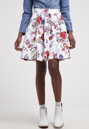 Kvinde iført et hvidt plisseret blomsterprintet skørt, blå skjorte med knapper, lysegrå sokker og hvide ankelstøvler på en ensfarvet baggrund.
