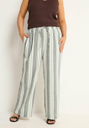 Personne portant un pantalon blanc ample à rayures verticales noires, un haut marron sans manches, et des sandales noires ouvertes à l'avant, debout avec les mains partiellement dans les poches.