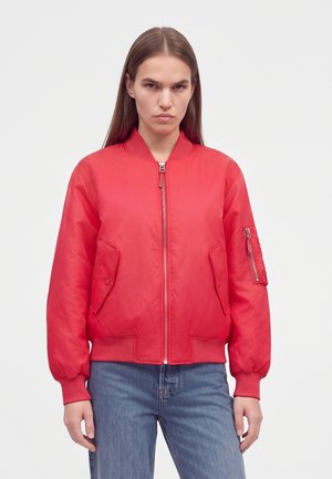 Jeune femme aux longs cheveux raides portant une veste bomber rouge et un jean bleu, debout devant un fond clair uni.