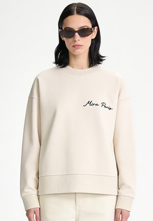 Frau mit schwarzen schulterlangen Haaren, die Schildpatt-Sonnenbrillen und einen cremefarbenen Pullover mit dem Text "Mira Paris" auf der Brust trägt.