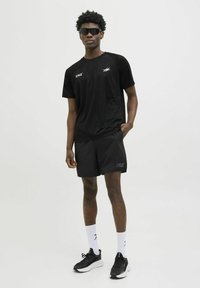 Sort atletisk t-shirt med logo på brystet, parret med sorte shorts, der har et logo. Båret med sorte sneakers og hvide sokker.