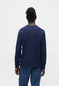 Tmavomodrá košeľa s dlhým rukávom z textúrovaného pleteného materiálu, s okrúhlym výstrihom a voľným strihom. Kombinovaná so svetlomodrými džínami z denimu.